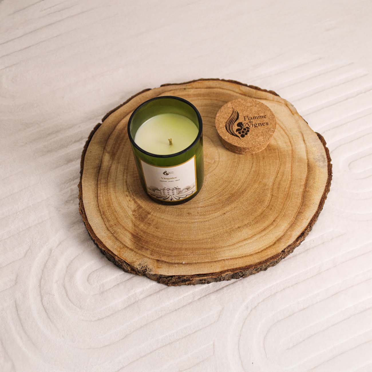 Bougie naturelle Viognier ouverte vue de dessus dans sa bouteille de vin recyclée verte, montrant la cire lisse et la mèche, avec son bouchon en liège gravé posé à côté sur un grand rondin de bois sur un tapis blanc texturé.