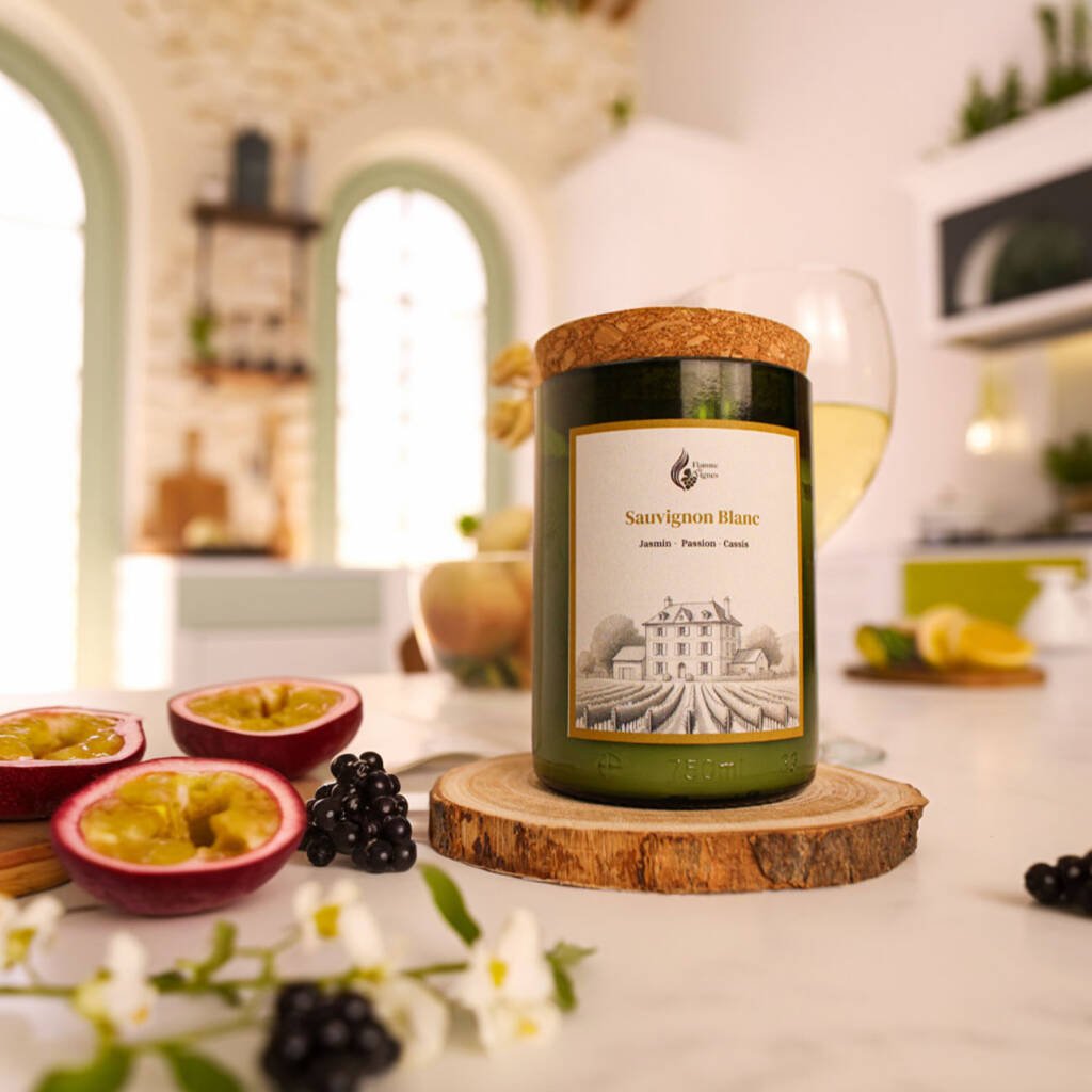 Bougie naturelle Sauvignon blanc en verre recyclé vert sur un socle en bois, entourée de fruits de la passion ouverts, de baies de cassis et de fleurs de jasmin dans une cuisine lumineuse aux fenêtres cintrées.