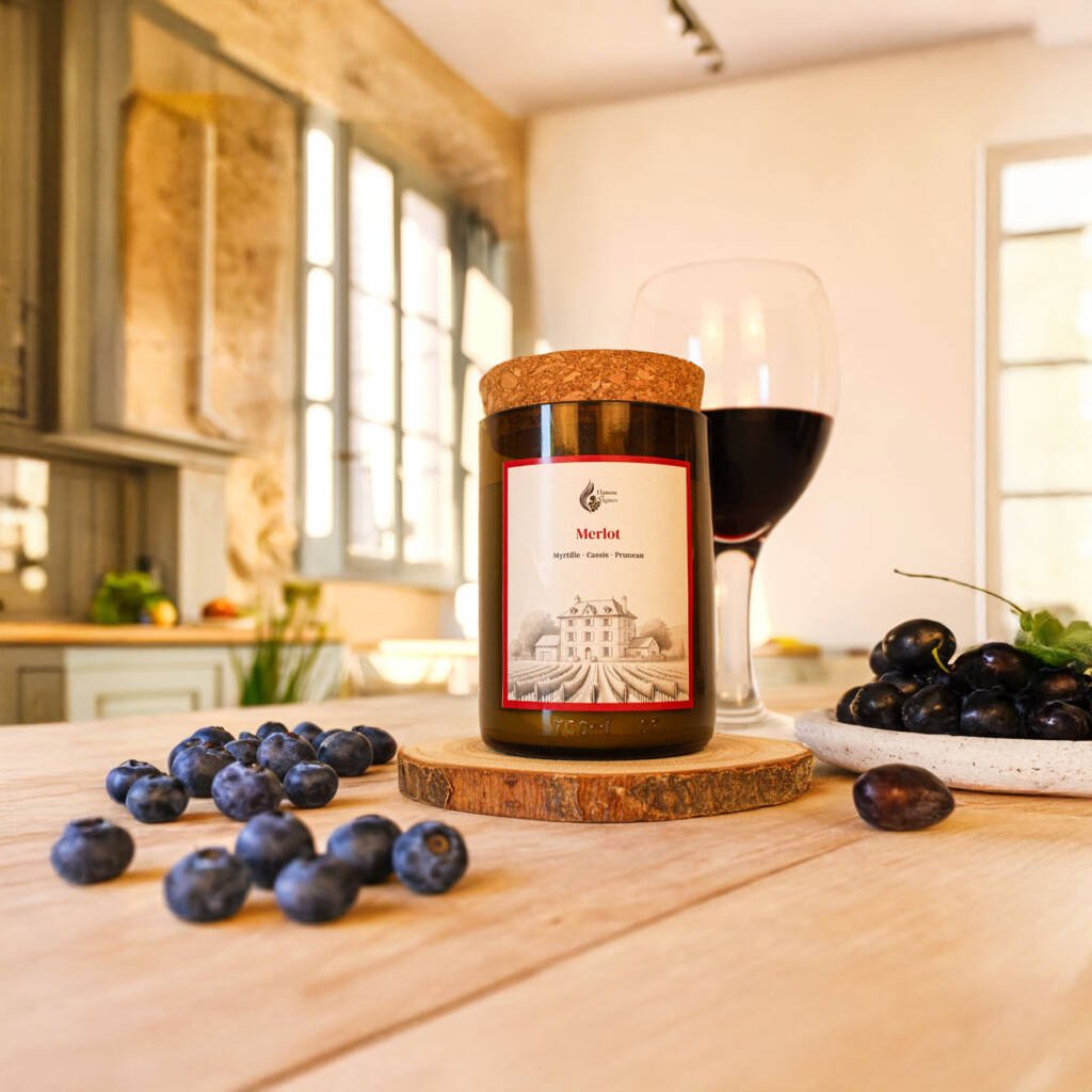 Bougie naturelle Merlot dans une authentique bouteille de vin recyclée sombre sur un rondin de bois, entourée de myrtilles fraîches éparpillées, d'une assiette de raisins noirs et d'un verre de vin rouge dans une cuisine lumineuse aux murs de pierre.