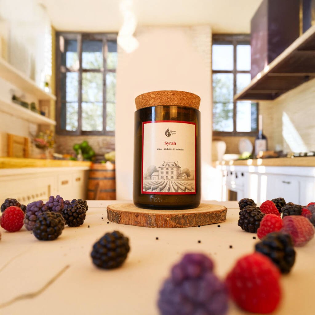 Bougie naturelle Syrah en verre recyclé sombre sur un rondin de bois, entourée de mûres et framboises fraîches éparpillées sur une table de cuisine lumineuse avec un tonneau en arrière-plan.